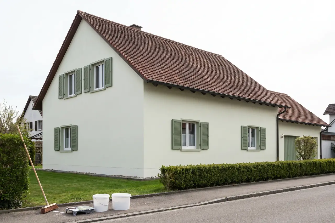 Dasselbe Wohnhaus frisch hell gestrichen, ohne Risse, grüne Fensterläden, Farbeimer am Gehweg